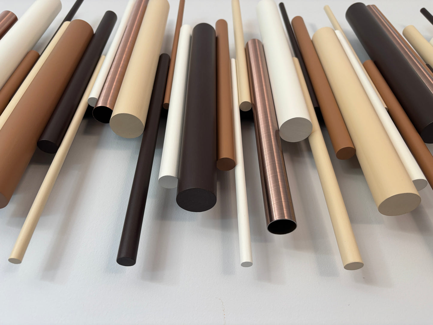 A collection of smooth, cylindrical rods or tubes in various matte and metallic finishes, displayed diagonally on a light gray background. The colors include shades of beige, brown, white, black, and antique copper tubes.