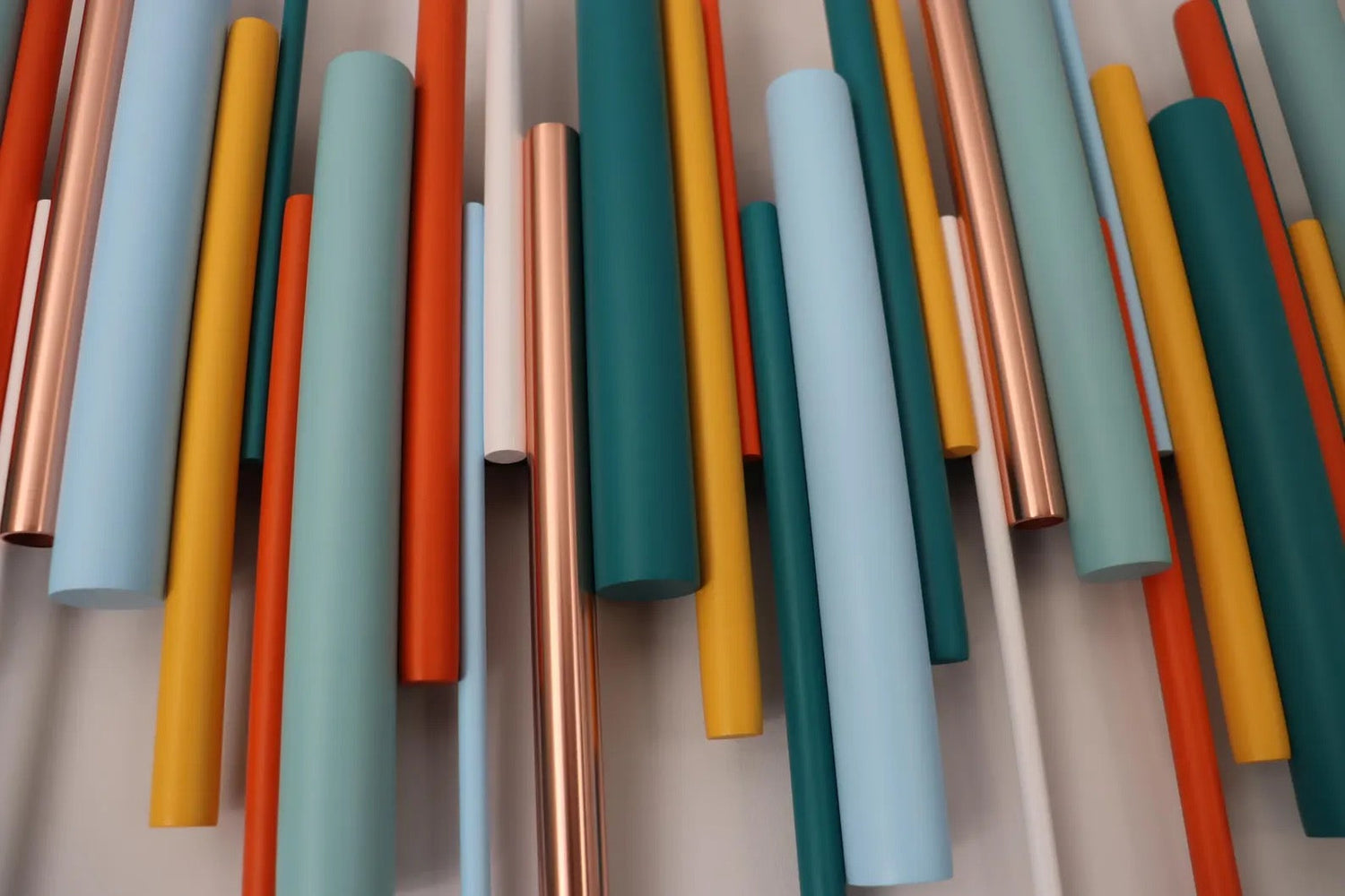 A close-up of multiple vertical cylindrical rods in matte and metallic finishes, including light blue, teal, orange, yellow, white, and polished copper, arranged against a light neutral background.