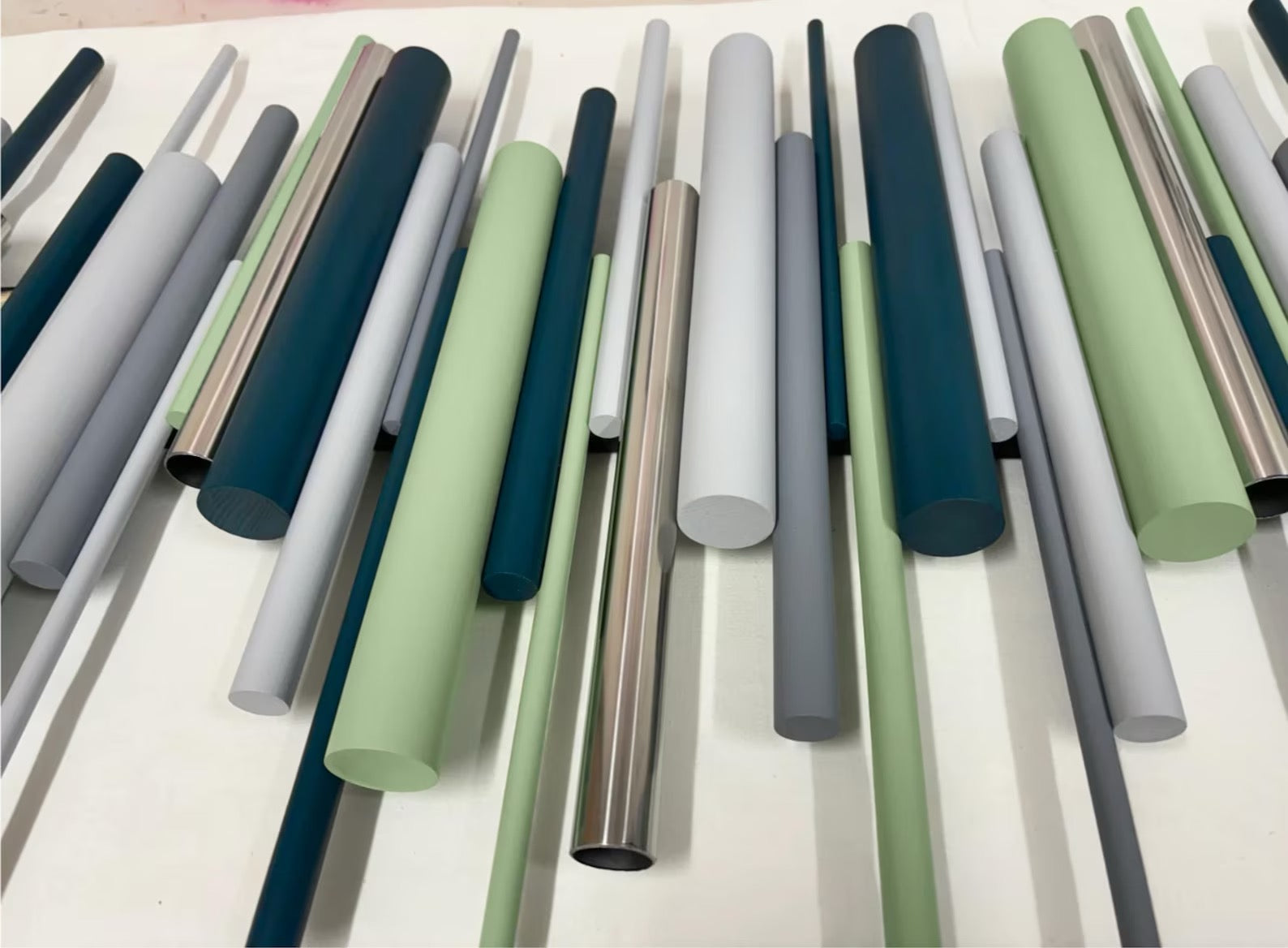 A collection of smooth, cylindrical rods or tubes in various matte and metallic finishes, displayed diagonally on a light surface. The colors include shades of sage green, slate blue, charcoal gray, light gray, and brushed silver.