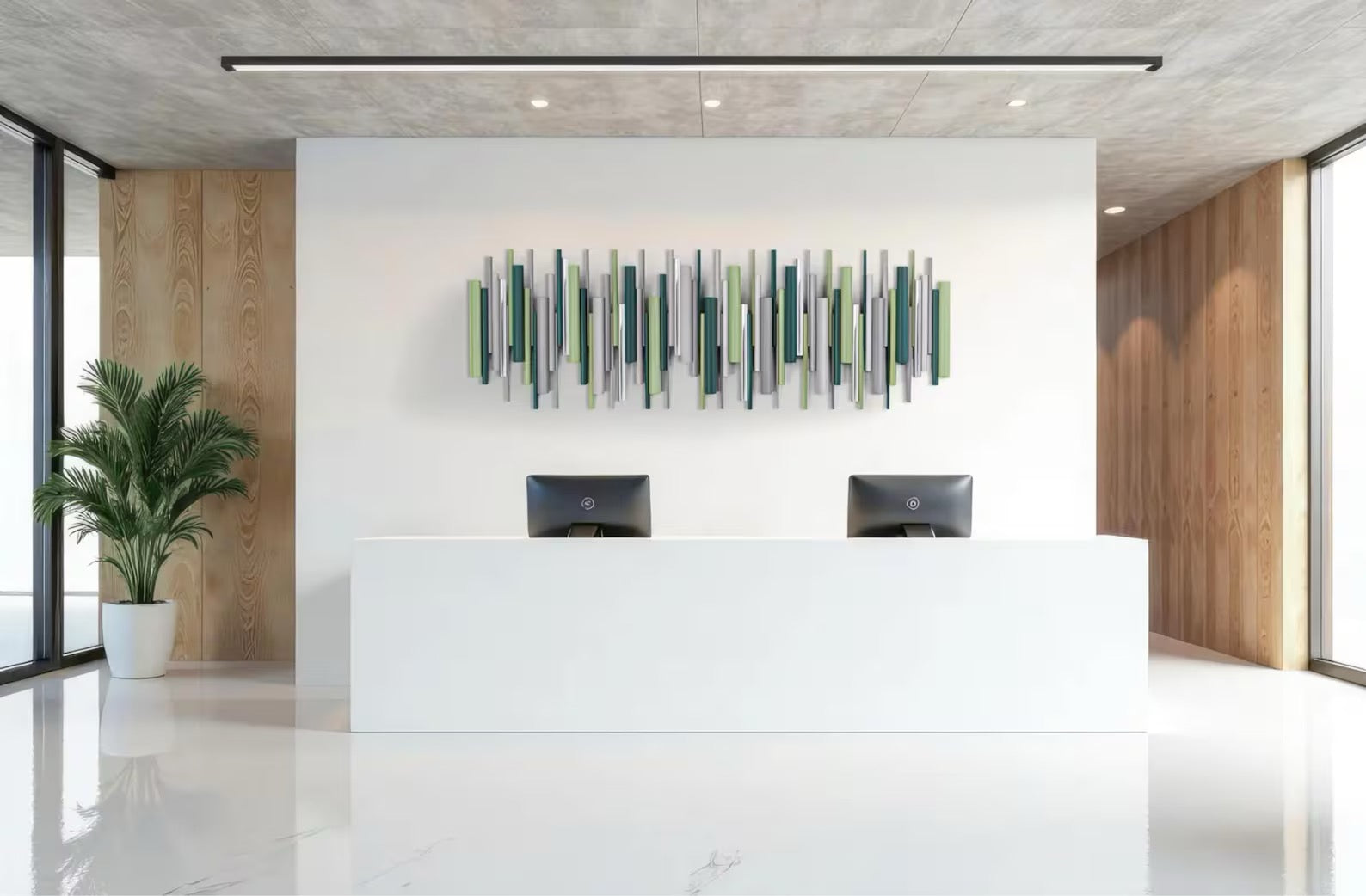 Modern reception desk in a bright office lobby with a white desk, two black computer monitors, a green potted plant, and a green and gray abstract wall art piece on a white wall.