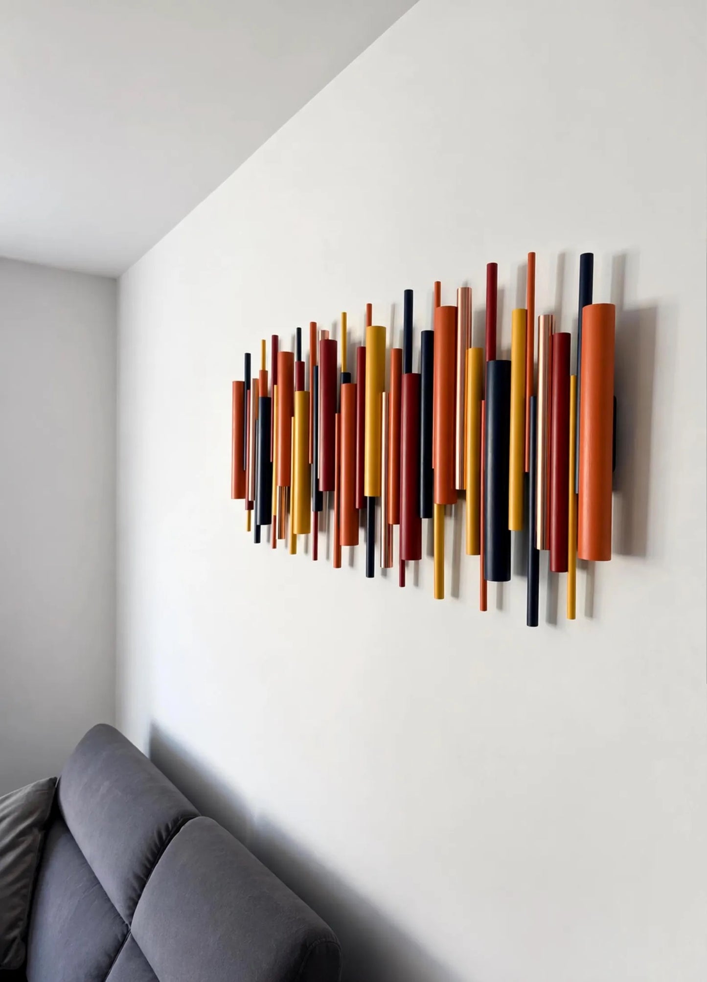 A modern wall art piece featuring multiple vertical wooden rods in orange, yellow, red, and black, mounted on a white wall above a gray sofa.
