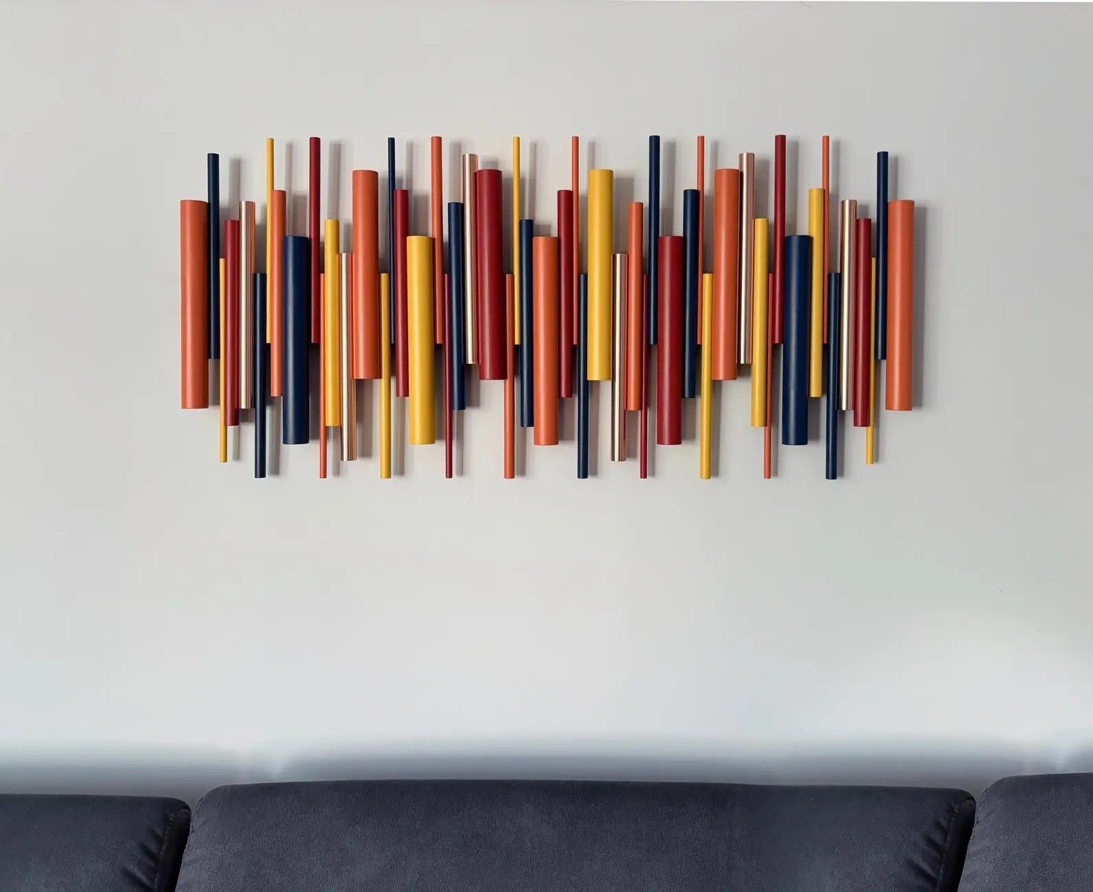 A modern wall-mounted art piece featuring multiple vertical cylindrical rods in orange, yellow, red, and navy blue, displayed on a light gray wall above a dark gray sofa.