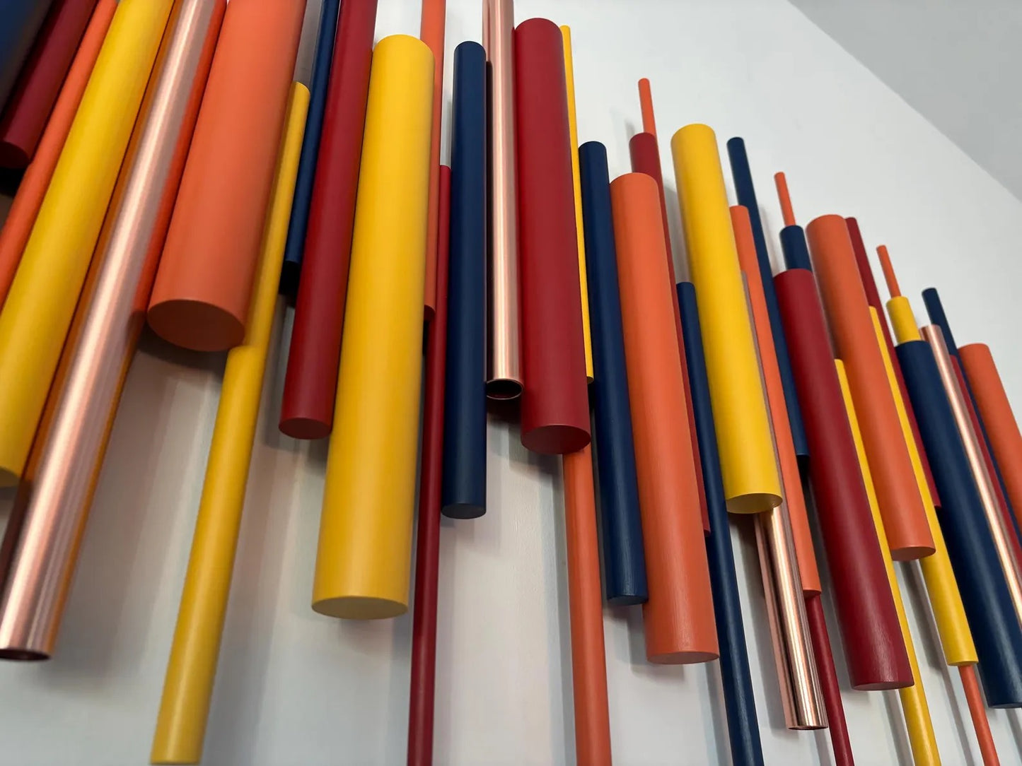 A wall-mounted abstract sculpture made of many vertical cylindrical rods in yellow, orange, red, navy, and metallic copper tones against a white wall.
