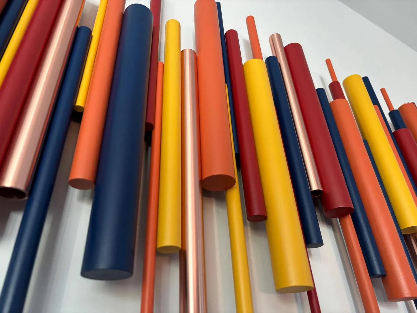 A collection of colorful cylindrical rods or tubes in yellow, orange, red, and navy blue, arranged vertically against a plain white wall.