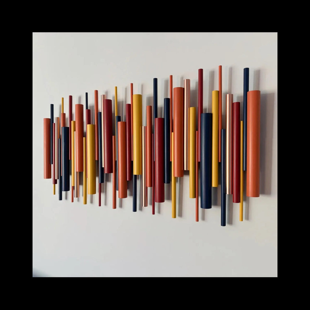 A modern wall-mounted art piece featuring multiple vertical cylindrical rods in rust red, mustard yellow, orange, and navy blue, arranged in a wavy, layered pattern against a plain white wall.