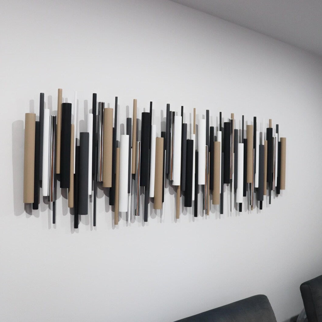 A modern wall-mounted art piece featuring vertical wooden slats in black, white, and natural beige tones, displayed on a plain white wall above a dark gray sofa.