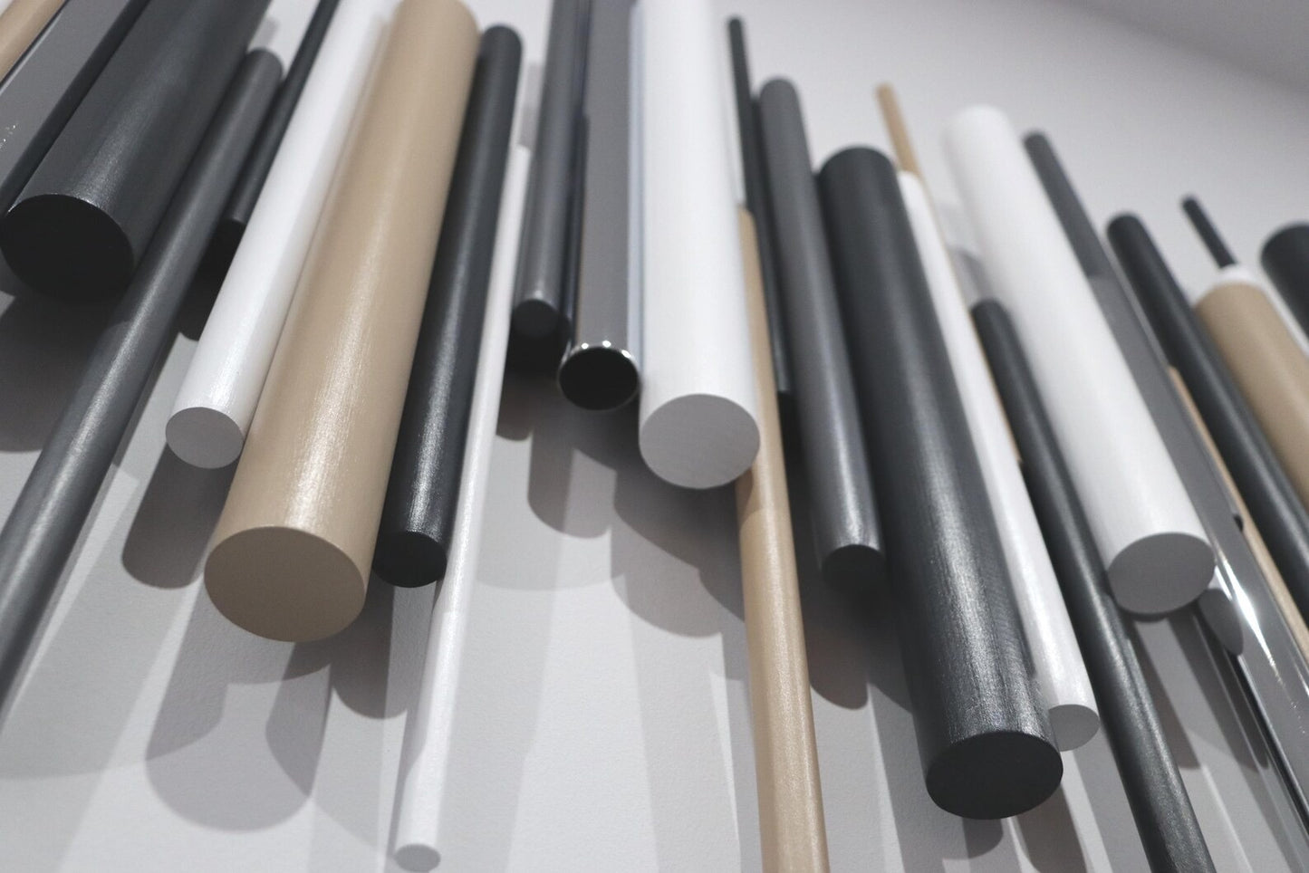 A close-up arrangement of cylindrical rods or dowels in matte black, glossy black, white, and warm beige/tan, photographed against a light neutral background.