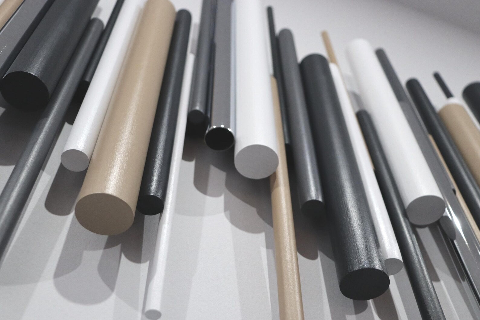 A close-up arrangement of cylindrical rods or dowels in matte black, glossy black, white, and warm beige/tan, photographed against a light neutral background.