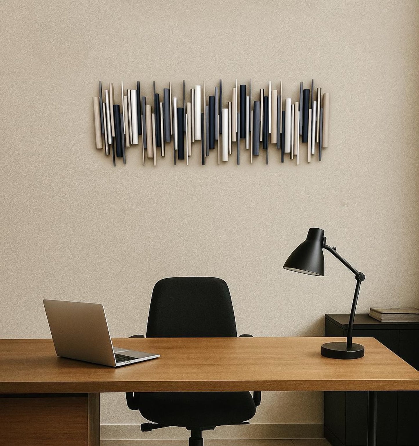 A modern wall-mounted art piece made of vertical metallic rods in silver, gray, and navy, displayed above a wooden desk with a black office chair, laptop, and black desk lamp.