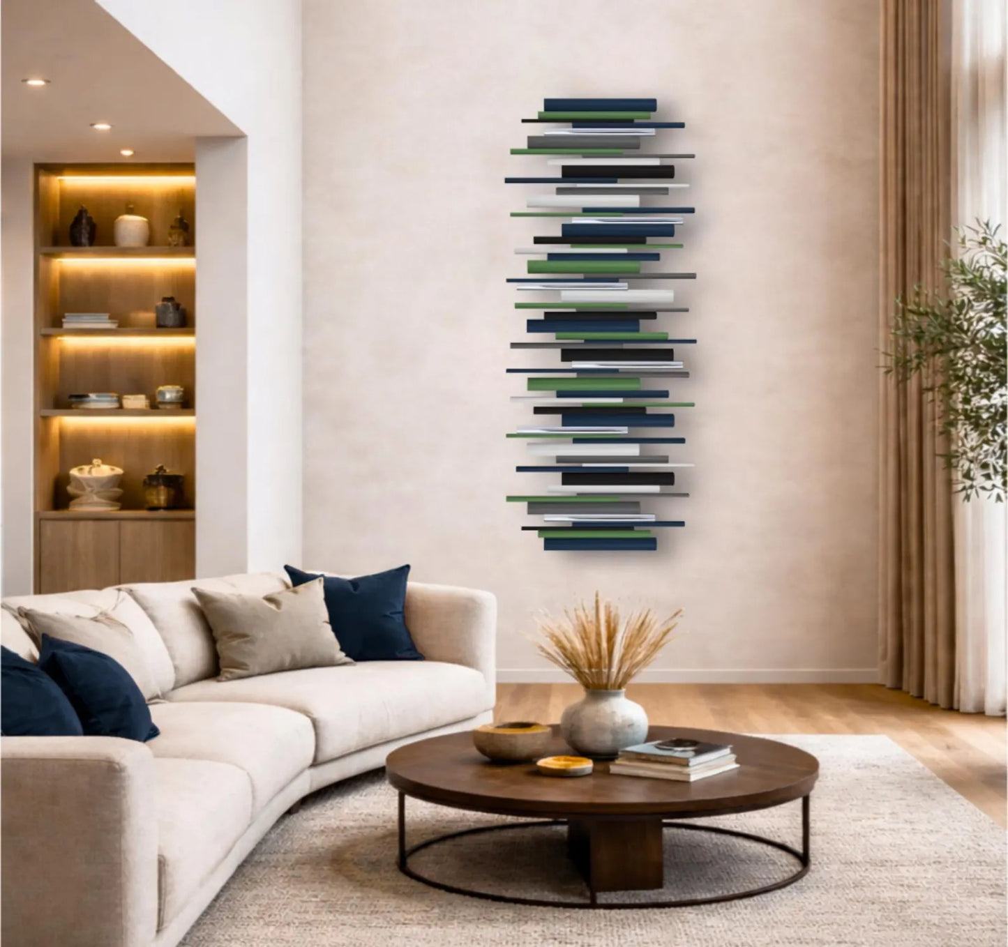 A modern curved white sofa in a warm beige living room, styled with navy and beige throw pillows, a round wooden coffee table, and a tall, narrow abstract wall art piece featuring horizontal painted wooden dowels in shades of navy, green, gray, and white.