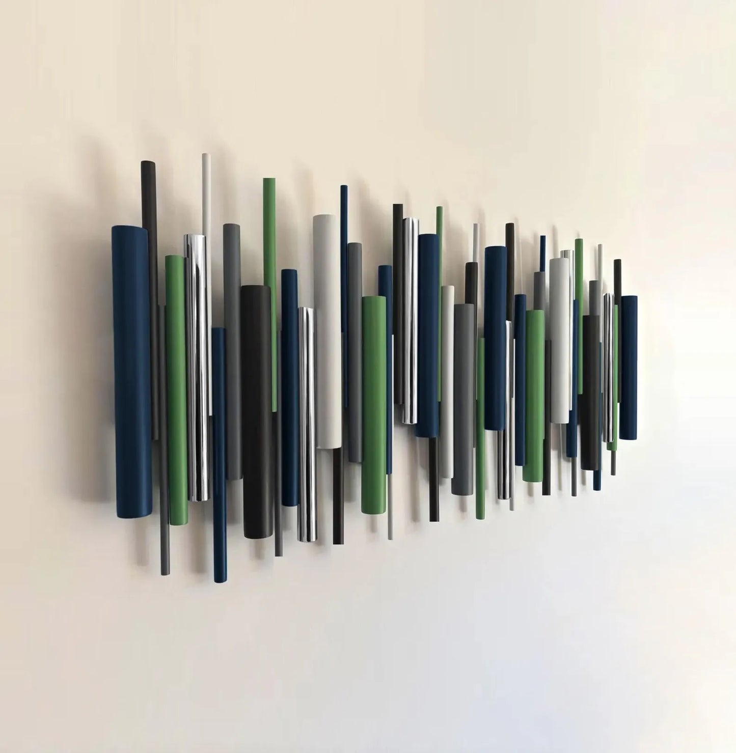 Abstract wall art featuring a series of vertical cylindrical rods in navy, green, gray, black, and white, mounted on a plain off-white wall.