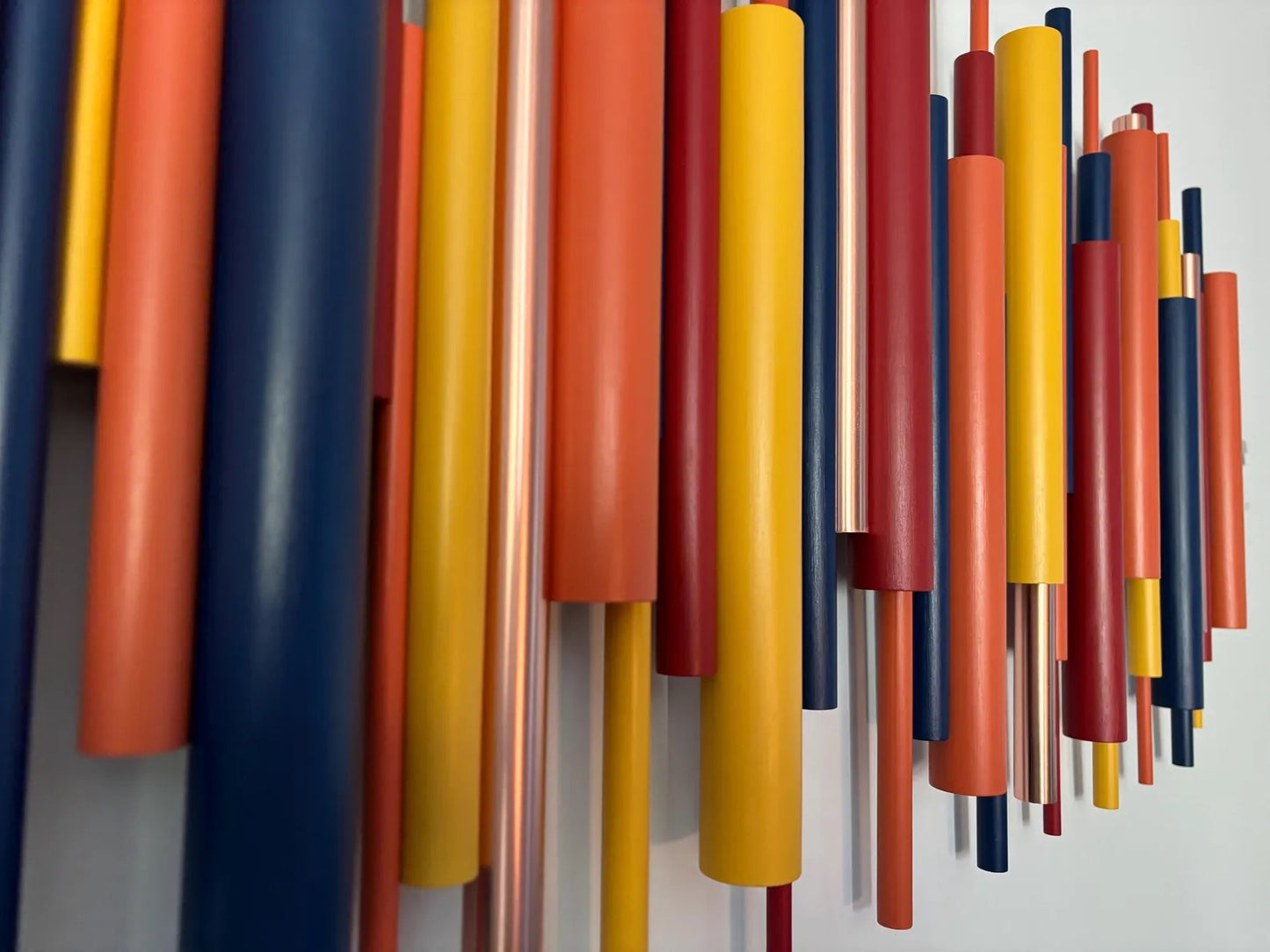 A close-up of a wall-mounted abstract art piece featuring multiple vertical cylindrical rods in matte orange, yellow, red, navy blue, and metallic copper tones against a light gray wall.