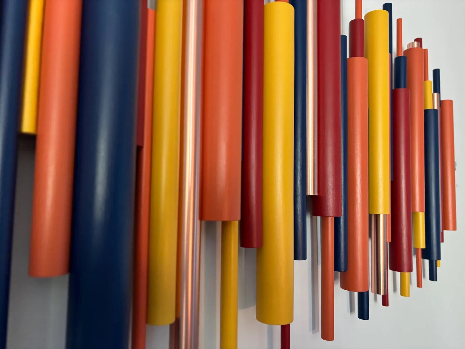 A close-up of a wall-mounted abstract art piece featuring multiple vertical cylindrical rods in matte orange, yellow, red, navy blue, and metallic copper tones against a light gray wall.