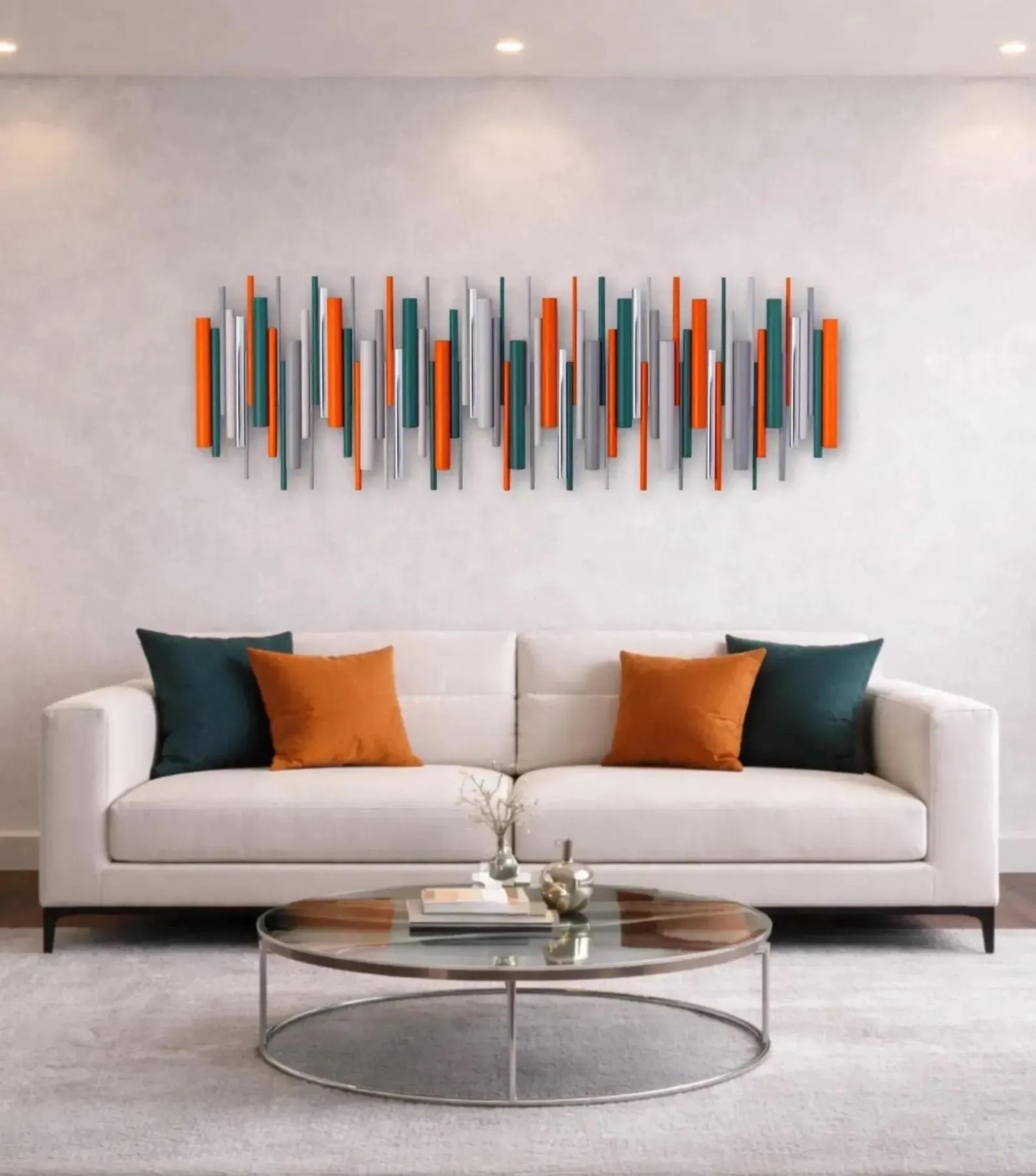 A  large abstract wood and metal wall art piece in orange, teal, and gray above the couch. Set in a living room scene featuring a white fabric sofa with dark teal and orange accent pillows, a round glass-top coffee table, and 